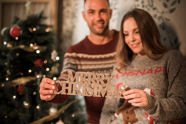 Homme souriant près de femme joyeuse avec tablette de joyeux Noël près de sapin décoré