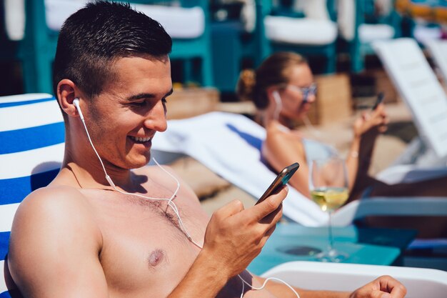 Homme souriant se détendre sur une chaise longue, en utilisant un téléphone portable, dans les écouteurs, écouter de la musique.