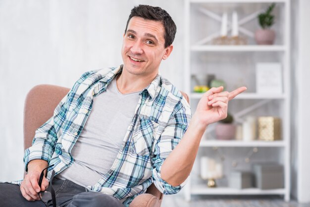 Homme souriant tenant des lunettes et pointant loin sur une chaise à la maison