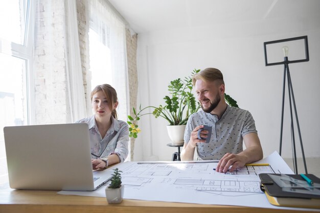 Homme souriant tenant une tasse de café en regardant un ordinateur portable en utilisant son collègue de travail au bureau