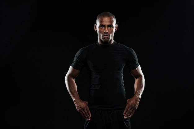 Homme de sport afro-américain sérieux avec les bras sur ses hanches en regardant la caméra