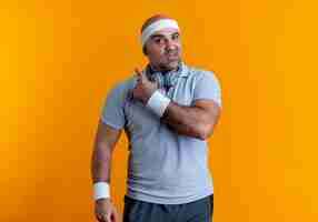 Photo gratuite homme sportif mature en bandeau à l'avant avec une expression confuse pointant vers l'arrière debout sur un mur orange