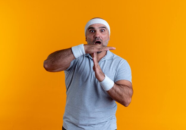 Homme sportif mature en bandeau à l'avant inquiet de faire le geste de temps avec les mains debout sur le mur orange