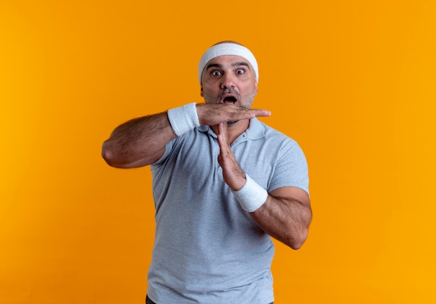 Homme sportif mature en bandeau à l'avant inquiet de faire le geste de temps avec les mains debout sur le mur orange