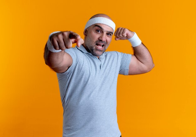 Homme sportif mature en bandeau à l'avant montrant les biceps pointant avec le doigt vers l'avant souriant debout sur le mur orange