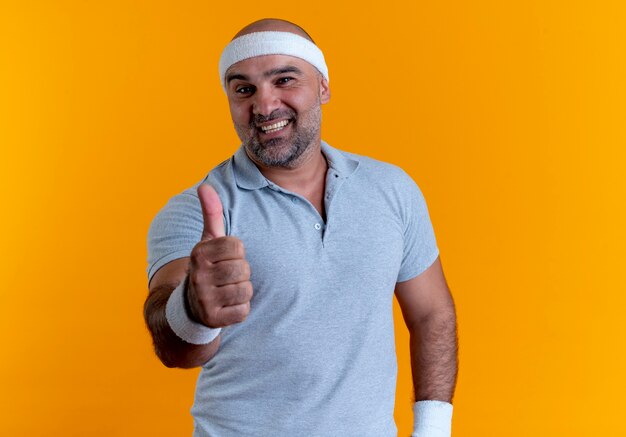 Homme sportif mature en bandeau à l'avant en souriant montrant les pouces vers le haut debout sur le mur orange