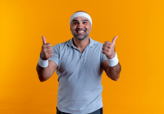 Homme sportif mature en bandeau à l'avant souriant avec visage heureux montrant les pouces vers le haut debout sur le mur orange