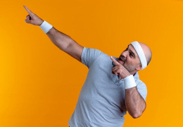 Homme sportif mature en bandeau à la confiance pointant avec les doigts sur le côté debout sur un mur orange