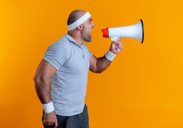 Homme sportif mature en bandeau criant au mégaphone debout sur un mur orange