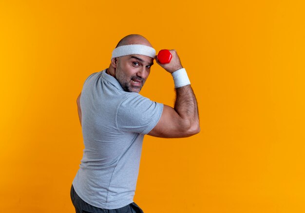 Homme sportif mature en bandeau main levée avec haltère à la tension et confiant debout sur le mur orange