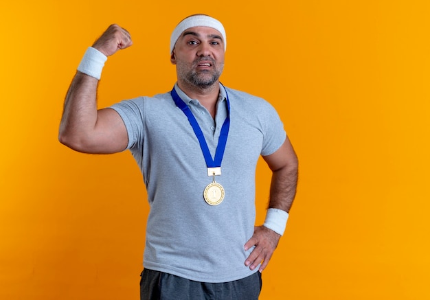 Homme sportif mature en bandeau avec médaille d'or autour de son cou levant le poing à la confiance debout sur le mur orange