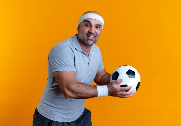 Homme sportif mature en bandeau tenant un ballon de football à l'avant avec le sourire sur le visage debout sur un mur orange