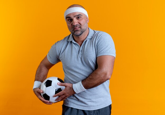 Homme sportif mature en bandeau tenant un ballon de football à l'avant avec un visage sérieux debout sur un mur orange