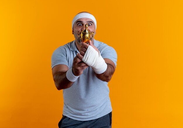 Homme sportif mature en bandeau tenant le trophée à la surprise et étonné debout sur le mur orange