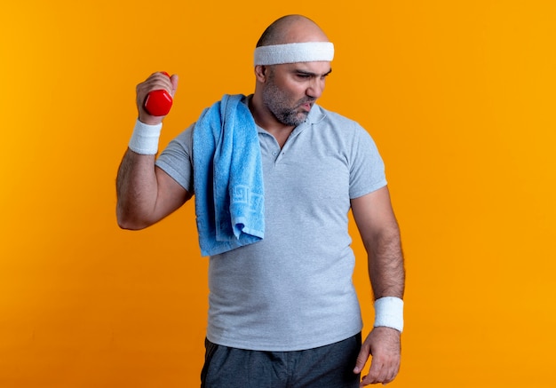 Homme sportif mature en bandeau travaillant avec haltère à la fatigue et épuisé debout sur le mur orange