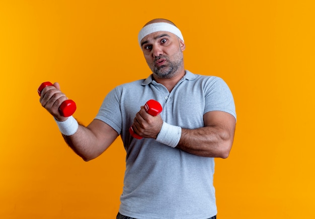 Homme sportif mature en bandeau travaillant avec des haltères à la confusion debout sur le mur orange