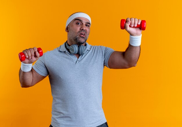 Homme sportif mature en bandeau travaillant avec des haltères à la tension et confiant debout sur le mur orange 3