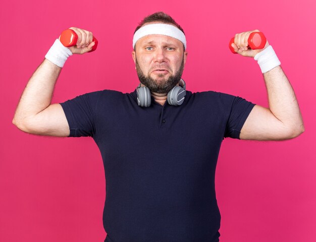 homme sportif slave adulte mécontent avec des écouteurs portant un bandeau et des bracelets tenant des haltères isolés sur un mur rose avec espace de copie