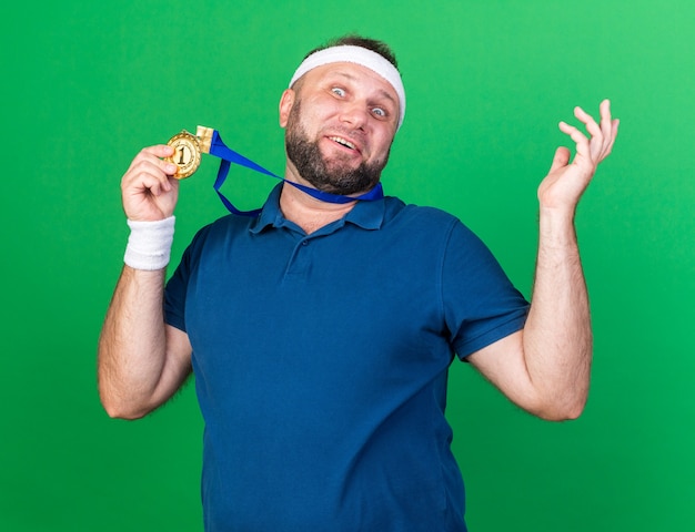 homme sportif slave adulte surpris portant un bandeau et des bracelets tenant une médaille d'or isolée sur un mur vert avec espace de copie