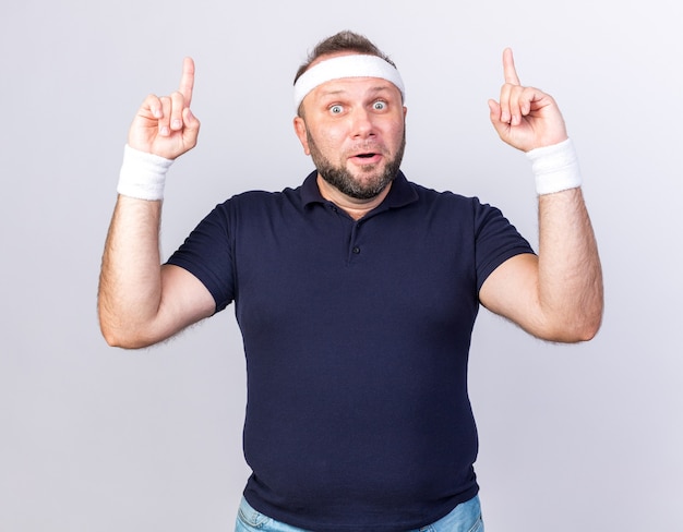 Homme sportif slave adultes surpris portant un bandeau et des bracelets pointant vers le haut isolé sur un mur blanc avec copie espace