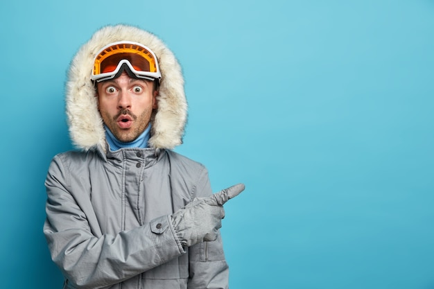 Photo gratuite un homme stupéfait actif s'entraîne dans les montagnes d'hiver, le snowboard porte une veste grise et des lunettes de ski indique avec une expression surprise sur un espace vide.