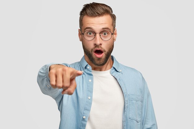 Homme stupéfait à la mode avec des poils, coupe de cheveux élégante, vêtu d'une veste en jean, vous montre avec une expression surprise, sélectionne quelque chose, isolé sur un mur blanc. Concept d'omg et de réaction.