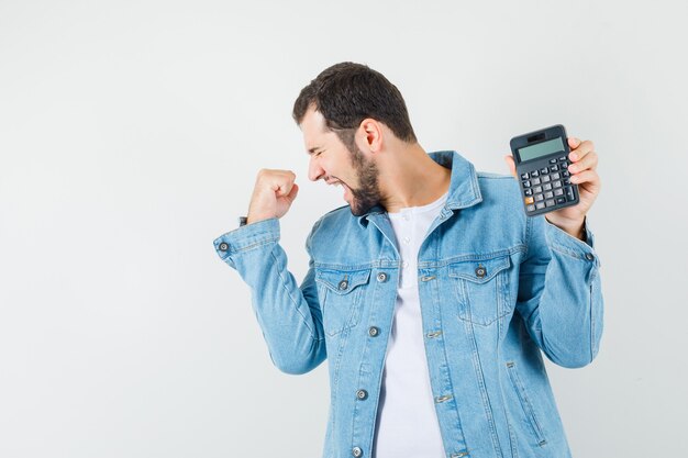 Homme de style rétro en veste, t-shirt montrant la calculatrice tout en montrant le geste du gagnant et en regardant heureux, vue de face. espace pour le texte