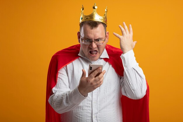 Homme de super-héros adultes agacé en cape rouge portant des lunettes et une couronne tenant et regardant le téléphone mobile en gardant la main dans l'air isolé sur un mur orange