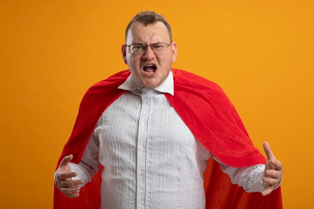 Homme de super-héros adultes en colère en cape rouge portant des lunettes en gardant les mains en l'air à l'avant isolé sur mur orange