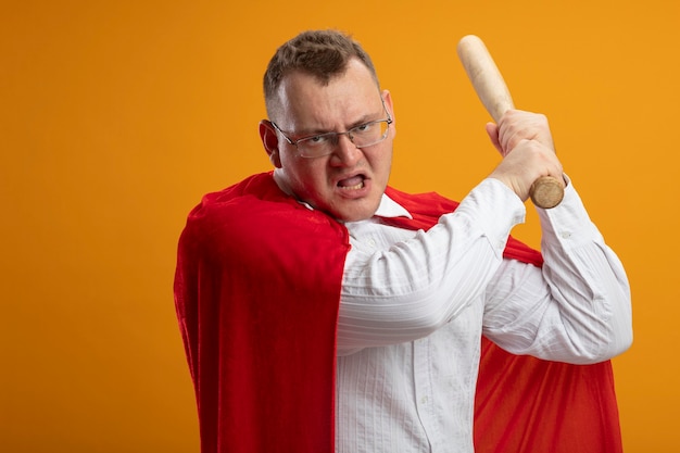 Homme de super-héros adultes en colère en cape rouge portant des lunettes tenant une batte de baseball à l'avant se prépare à frapper isolé sur mur orange