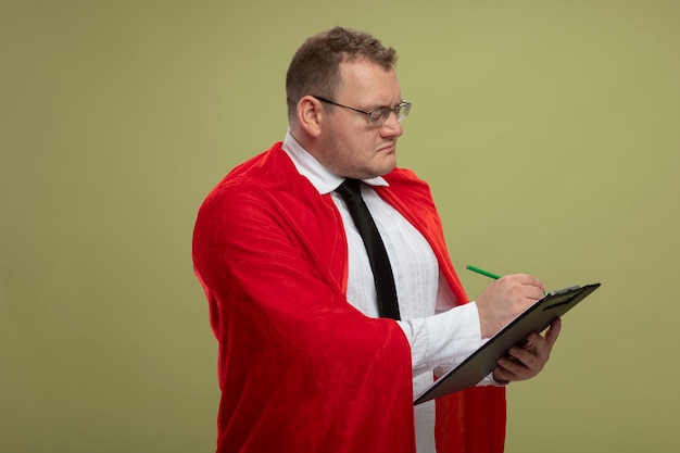 Homme de super-héros adultes concentré en cape rouge portant des lunettes debout en vue de profil écrit sur le presse-papiers avec un stylo isolé sur mur vert olive