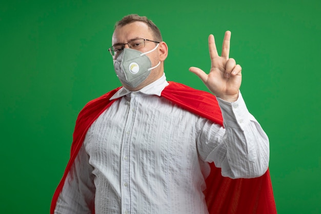 Homme de super-héros adultes confiant en cape rouge portant des lunettes et un masque de protection à l'avant montrant trois avec main isolé sur mur vert