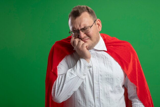 Homme de super-héros adultes confus en cape rouge portant des lunettes mettant la main sur le visage à l'avant isolé sur mur vert