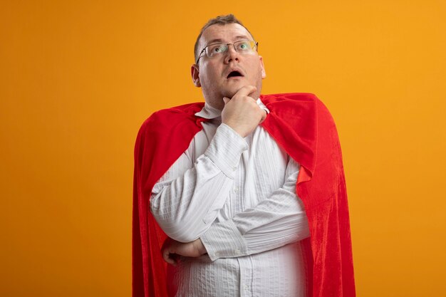 Homme de super-héros adultes impressionné en cape rouge portant des lunettes touchant le menton en levant isolé sur mur orange