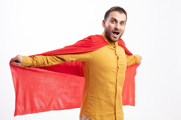Homme de super-héros caucasien excité se tient en gardant les bras ouverts tenant un manteau rouge isolé sur un mur blanc avec espace de copie