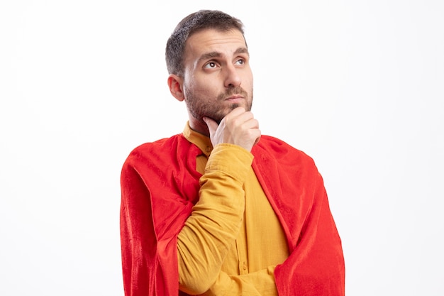 Homme de super-héros réfléchi avec manteau rouge met la main sur le menton et regarde à côté isolé sur mur blanc