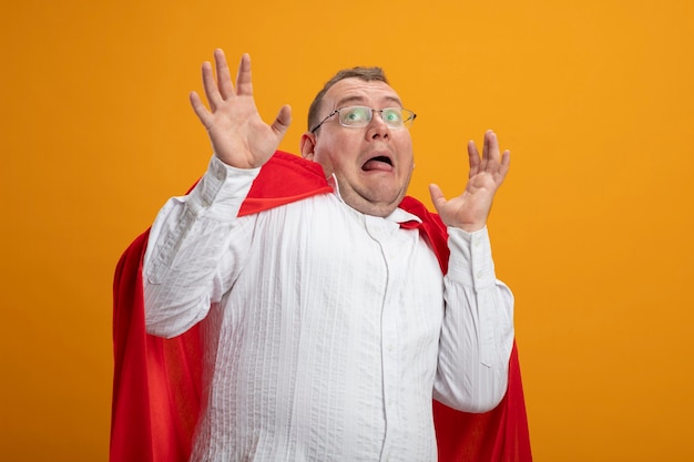 Homme de super-héros slaves adultes effrayés en cape rouge portant des lunettes en gardant les mains en l'air en levant isolé sur fond orange avec copie espace