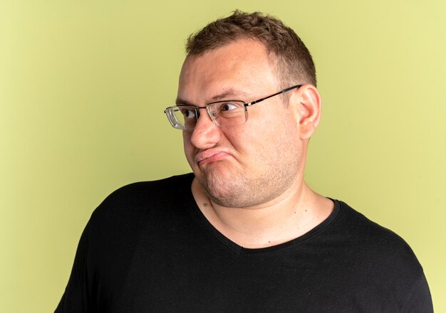 L'homme en surpoids dans des verres portant un t-shirt noir à côté mécontent de faire la grimace debout sur un mur léger