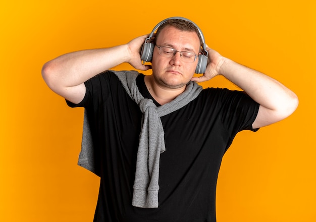 Homme en surpoids dans des verres portant un t-shirt noir avec des écouteurs bénéficiant de la musique debout sur un mur orange