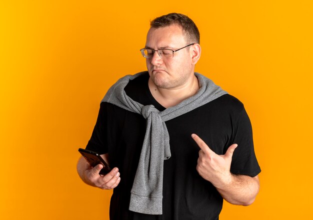 L'homme en surpoids dans des verres portant un t-shirt noir holding smartphone à mécontent montrant l'index debout sur le mur orange