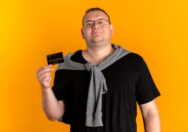 Homme en surpoids dans des verres portant un t-shirt noir montrant une carte de crédit regardant la caméra avec le sourire sur le visage debout sur un mur orange