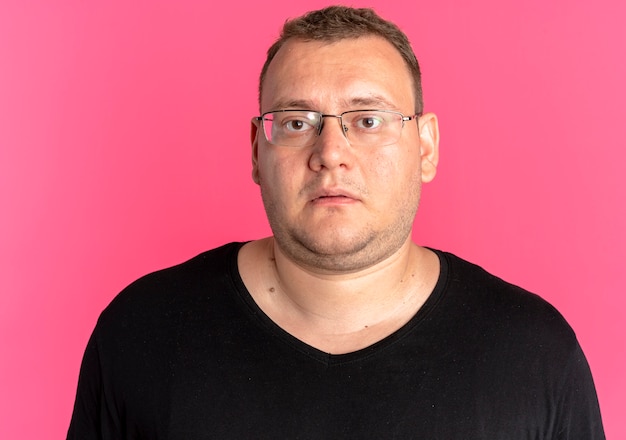 Homme en surpoids dans des verres portant un t-shirt noir regardant la caméra avec une expression triste debout sur un mur rose