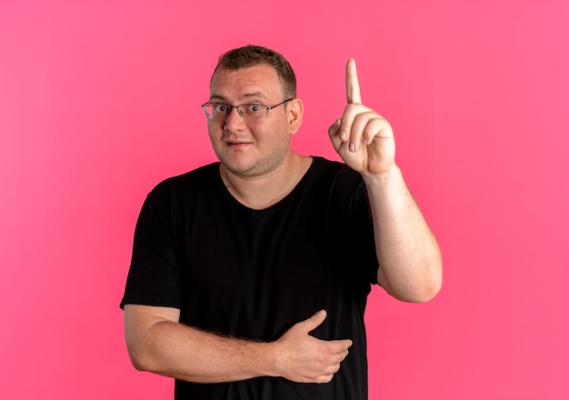 Homme en surpoids dans des verres portant un t-shirt noir souriant montrant l'index ayant une nouvelle excellente idée debout sur un mur rose