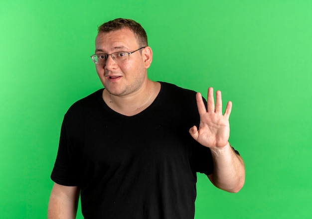 Homme en surpoids dans des verres portant un t-shirt noir à la surprise en agitant avec la main debout sur le mur vert