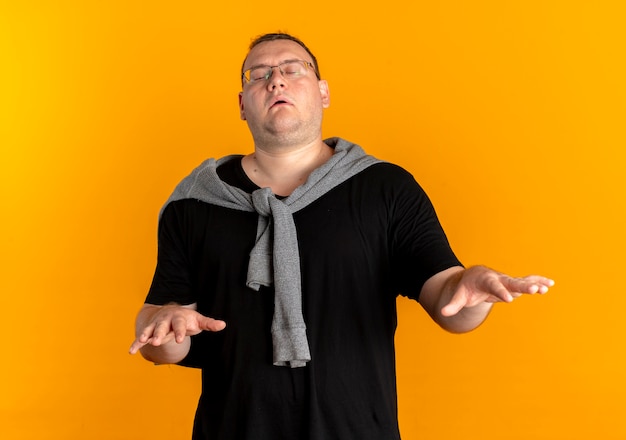 Homme en surpoids dans des verres portant un t-shirt noir tenant les mains avec les yeux fermés debout sur le mur orange