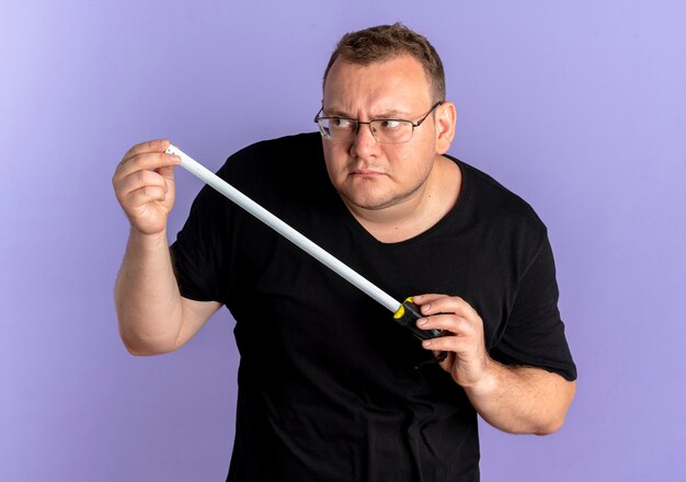 Homme en surpoids dans des verres portant un t-shirt noir tenant une règle à côté comme un espion debout sur un mur bleu