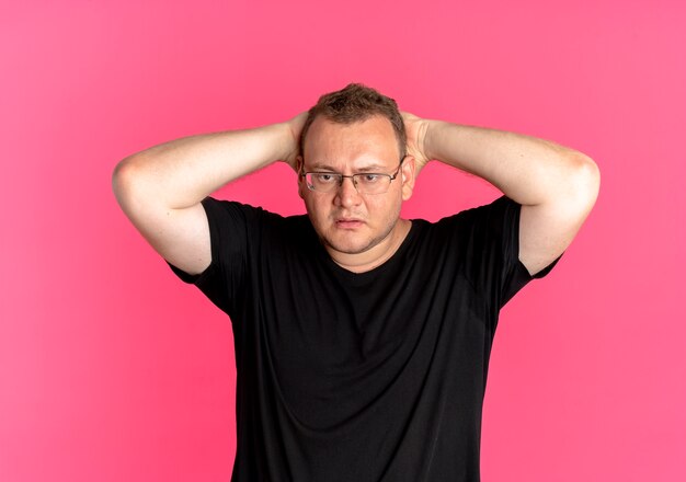 Homme en surpoids à lunettes portant un t-shirt noir à la confusion avec les mains sur sa tête sur rose