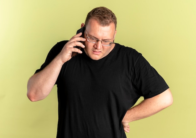 Homme En Surpoids à Lunettes Portant Un T-shirt Noir à La Confusion Et Mécontent Tout En Parlant Au Téléphone Mobile Debout Sur Un Mur Léger