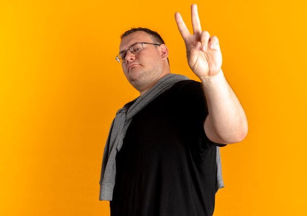 Homme en surpoids à lunettes portant un t-shirt noir à la recherche de confiance montrant signe de la victoire debout sur un mur orange