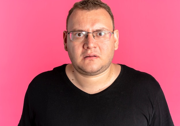 Homme en surpoids à lunettes portant un t-shirt noir regardant la caméra surpris et mécontent debout sur le mur rose